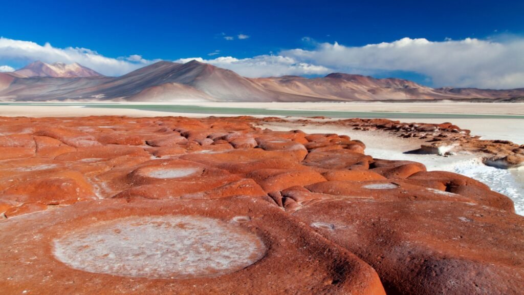 Piedras rojas e salar de água calientes no Atacama com vulcões ao fundo.